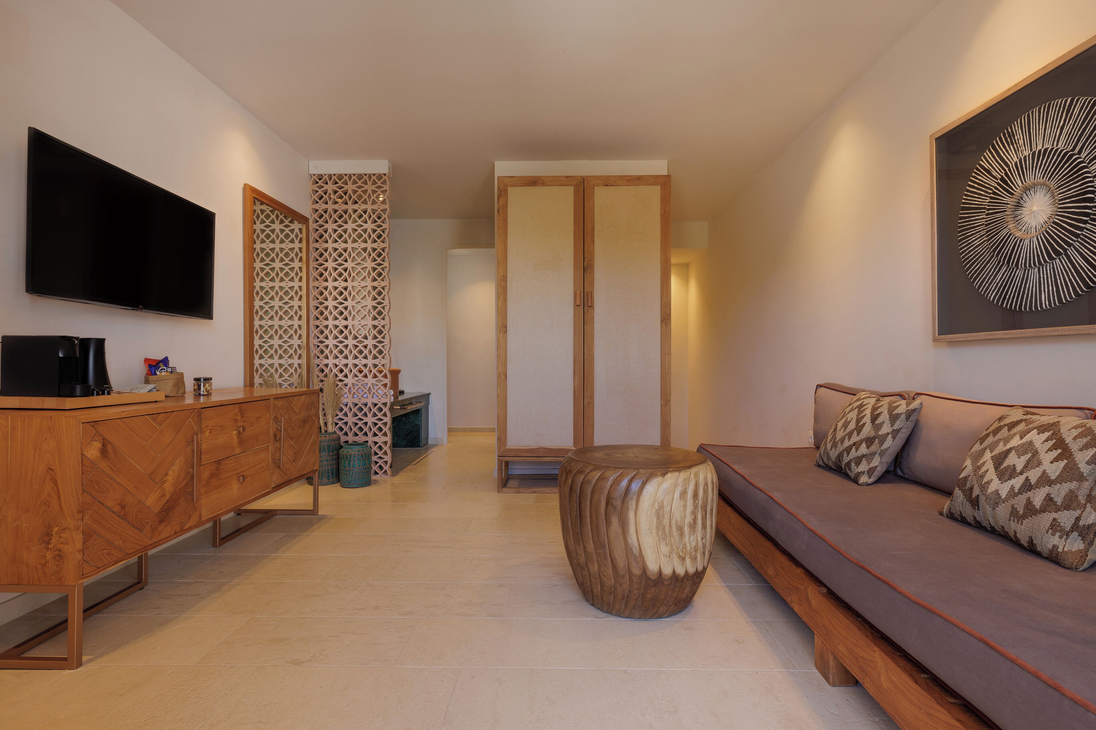Een moderne woonkamer met een houten kast, een rond houten krukje, een bank met opvallende kussens, een flatscreen televisie en een decoratief wandobject.