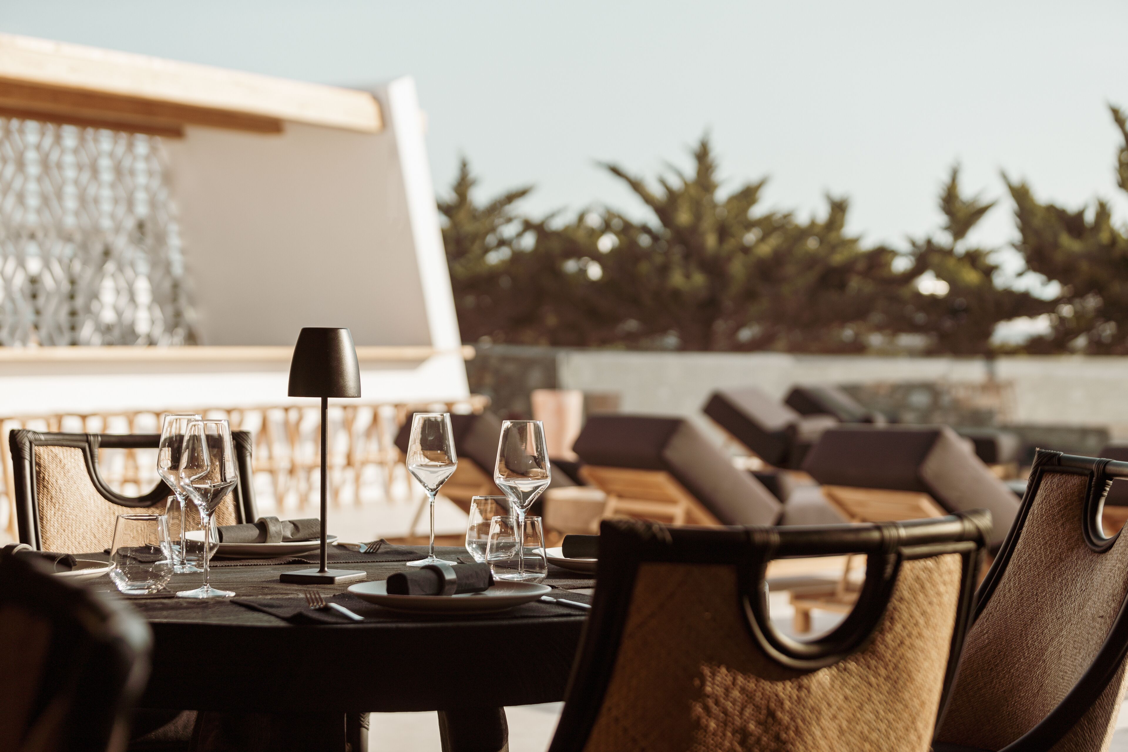 A dining table set with wine glasses, plates, and cutlery, surrounded by lounge chairs and a view of trees.