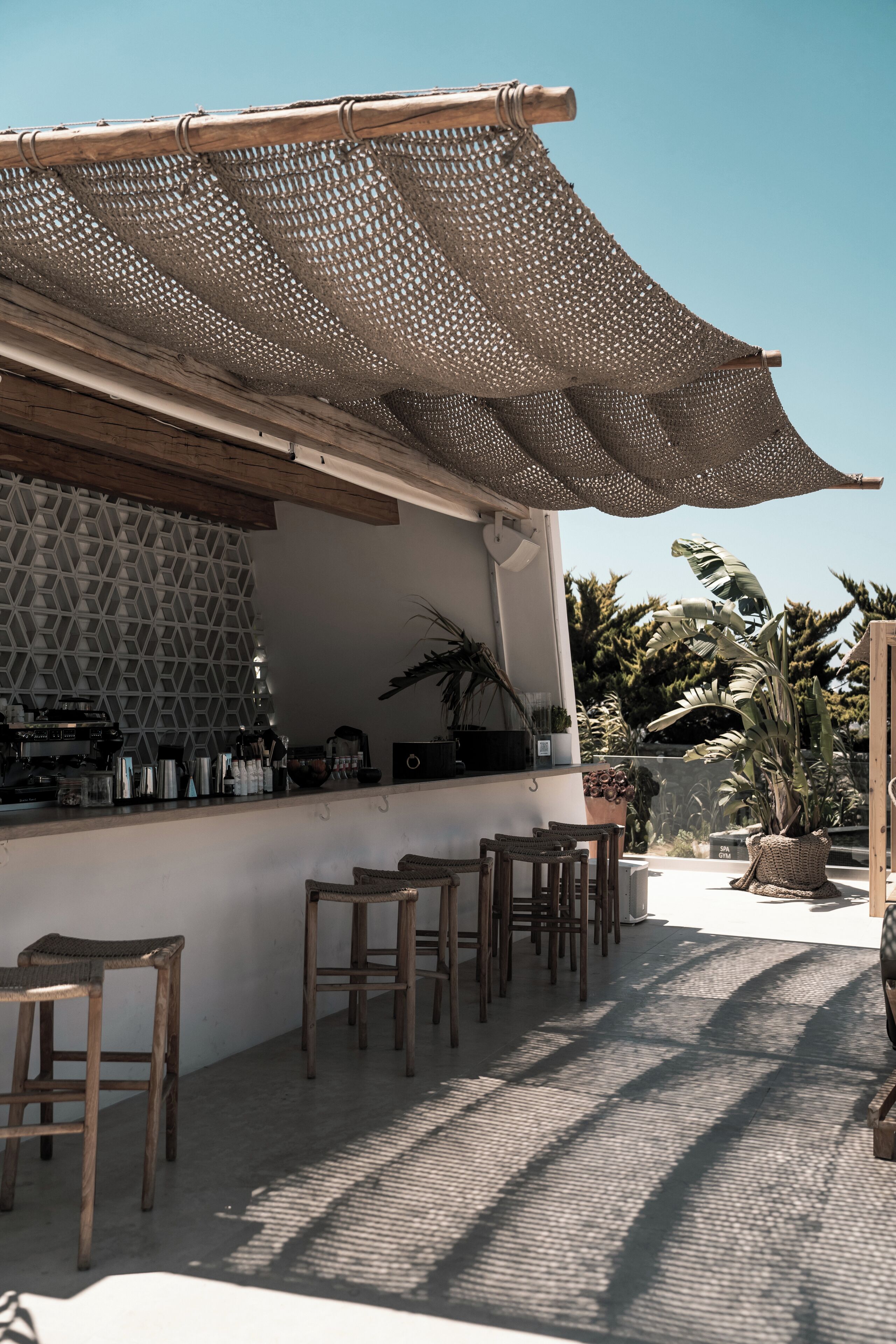 Ein Außenbereich mit Bar, der von einer Bambus-Markise überspannt ist, mit Holzstühlen und einer Wand mit Musterdesign.