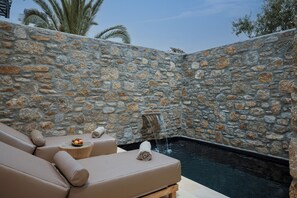 Un espacio al aire libre con paredes de piedra, un jacuzzi, sillones de descanso y una mesa de madera con fruta.