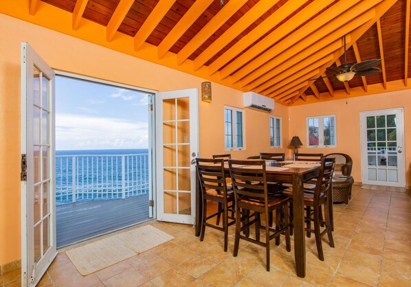 A dining area with a large table and chairs, a view of the ocean through an open door, and a ceiling with wooden beams.