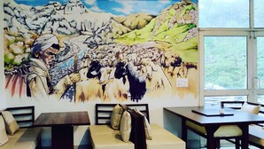 A room with a large mural of a mountainous landscape and a person leading a herd of animals. There are several tables and chairs arranged for dining. - Manuni Inn - Riverside Hotel (Dharamshala)