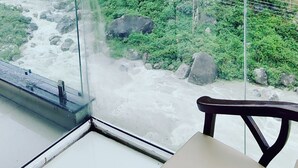 A glass-enclosed room with a view of a mountainous landscape, a chair, and a table. - Manuni Inn - Riverside Hotel (Dharamshala)