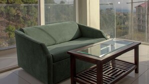 A green sofa and a glass-top table in a room with large windows. - Manuni Inn - Riverside Hotel (Dharamshala)
