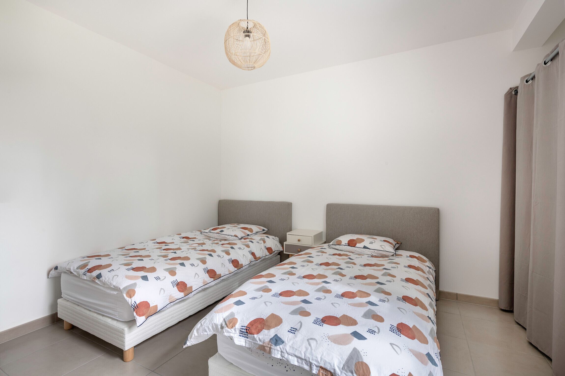 A bedroom with two single beds, a bedside table, a hanging light fixture, and a curtain on the right side.