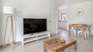 Una sala moderna con un televisor de pantalla plana sobre un soporte blanco, una mesa de centro de madera, una mesa de comedor con sillas blancas y una lámpara blanca.