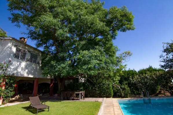 A pool area with a clear blue pool, a lounge chair, and a patio with a table and chairs.