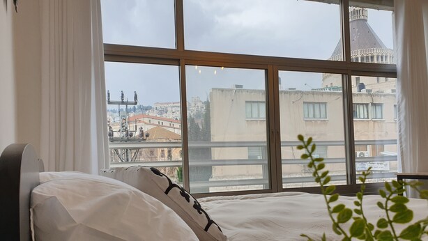 A bedroom with a large window, a bed with white bedding, and a view of buildings and a tower outside. - City Center Rooms (Nazareth)