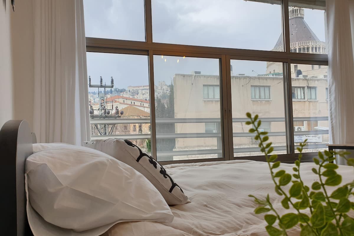 A bedroom with a large window, a bed with white bedding, and a view of buildings and a tower outside.