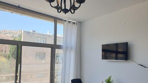A hotel room with a large window, a desk with a chair, a TV mounted on the wall, and a potted plant on the desk. - City Center Rooms (Nazareth)