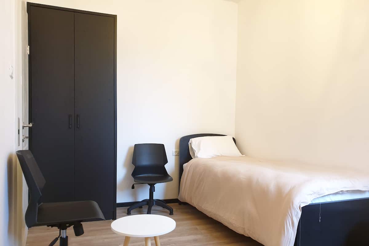 A small, neatly arranged bedroom with a bed, a chair, a small table, and a wardrobe.