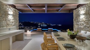 A dining area with a view of a city at night, stone walls, and wooden furniture. - Untold Mykonos (Mykonos)