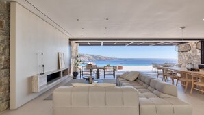 A modern living room with a large beige sectional sofa, a stone accent wall, and a view of the sea. - Untold Mykonos (Mykonos)