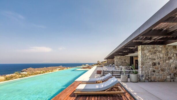 A luxurious outdoor infinity pool with wooden decking, lounge chairs, and a stone wall. - Untold Mykonos (Mykonos)
