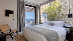 A modern bedroom with a large bed, a view of the sea, a stone wall, and a hanging light fixture. - Untold Mykonos (Mykonos)