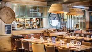 Ein Restaurantinneres mit Holztischen, Stühlen, einer großen Uhr und einer Theke, an der ein Koch Speisen zubereitet.