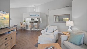 Una sala de estar moderna con un sofá, un reposapiés y una mesa de centro. Se observa un comedor con sillas y una mesa, y una cocina al fondo.