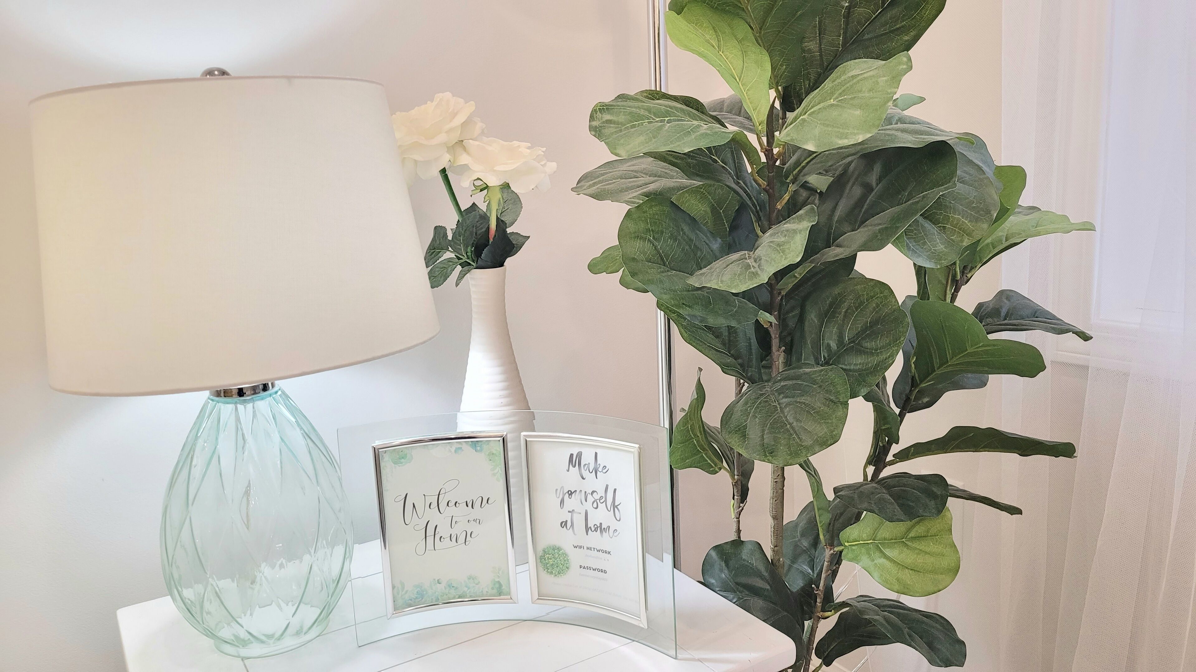 Une table avec une lampe, un vase de fleurs blanches et des panneaux encadrés sur lesquels on peut lire : « Bienvenue chez nous » et « Faites comme chez vous ».