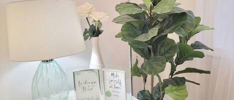 Une table avec une lampe, un vase de fleurs blanches et des panneaux encadrés sur lesquels on peut lire : « Bienvenue chez nous » et « Faites comme chez vous ».
