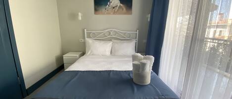 A neatly made bed with a blue bedspread and white pillows, a white bedside table, a wall-mounted air conditioner, and a painting of a horse.