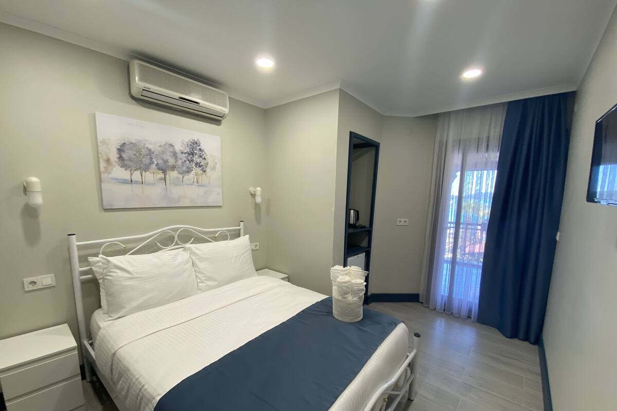 A bedroom with a bed, bedside tables, an air conditioner, a painting, a window with curtains, and a TV.