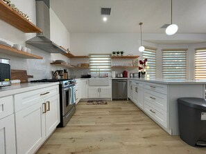 Una cuina moderna amb armaris blancs, electrodomèstics d'acer inoxidable, prestatges de fusta i una illa central.