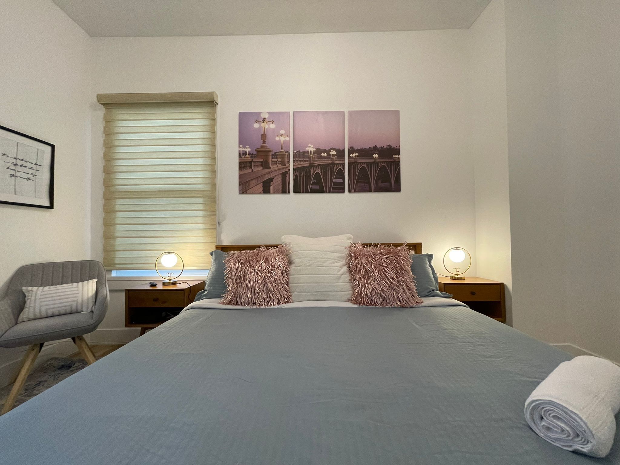 Un dormitorio con cama, dos mesitas de noche con lámparas, una silla, una ventana con persianas y una pared con tres cuadros enmarcados.