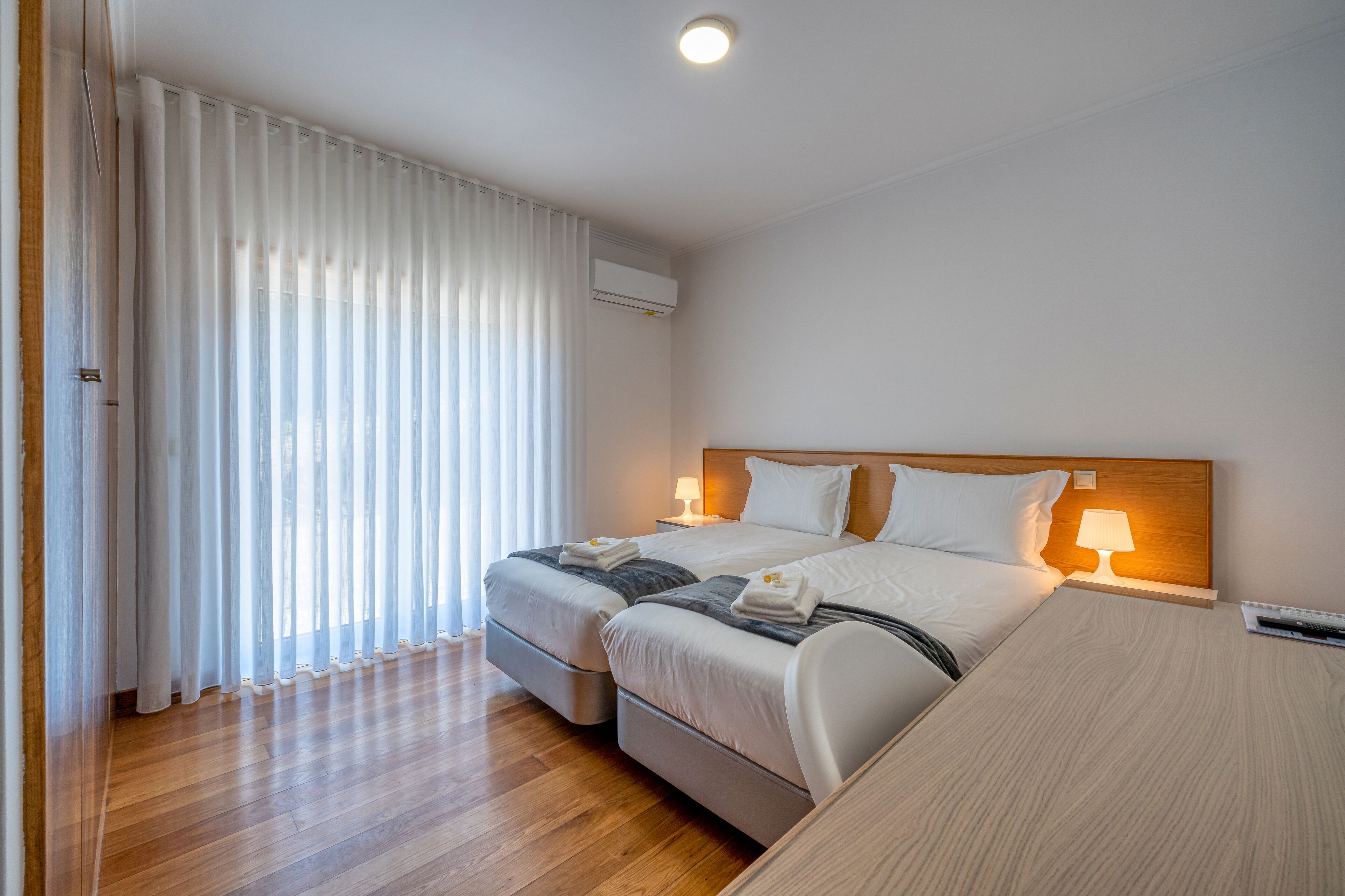 Habitación de hotel con dos camas, suelo de madera, un ventanal grande con cortinas translúcidas, equipo de aire acondicionado y lámparas de noche.