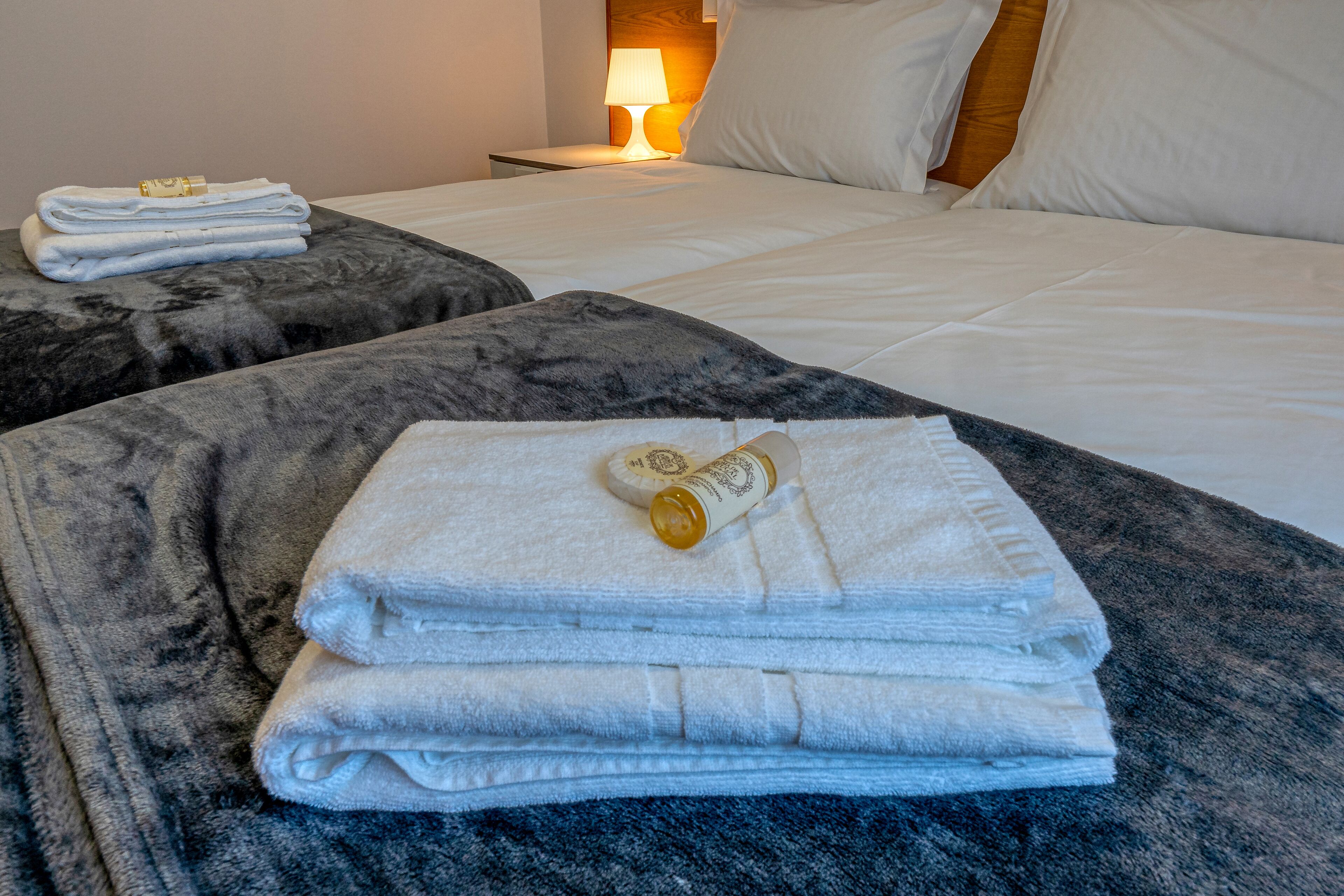 A neatly made bed with white linens and a rolled towel on a dark, textured ottoman.