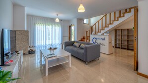 A modern living room with a grey sofa, a white coffee table, and a TV on a stand. There is a staircase leading to another level and a sign that reads "Central Suites". - Central Suites Arouca (Arouca)