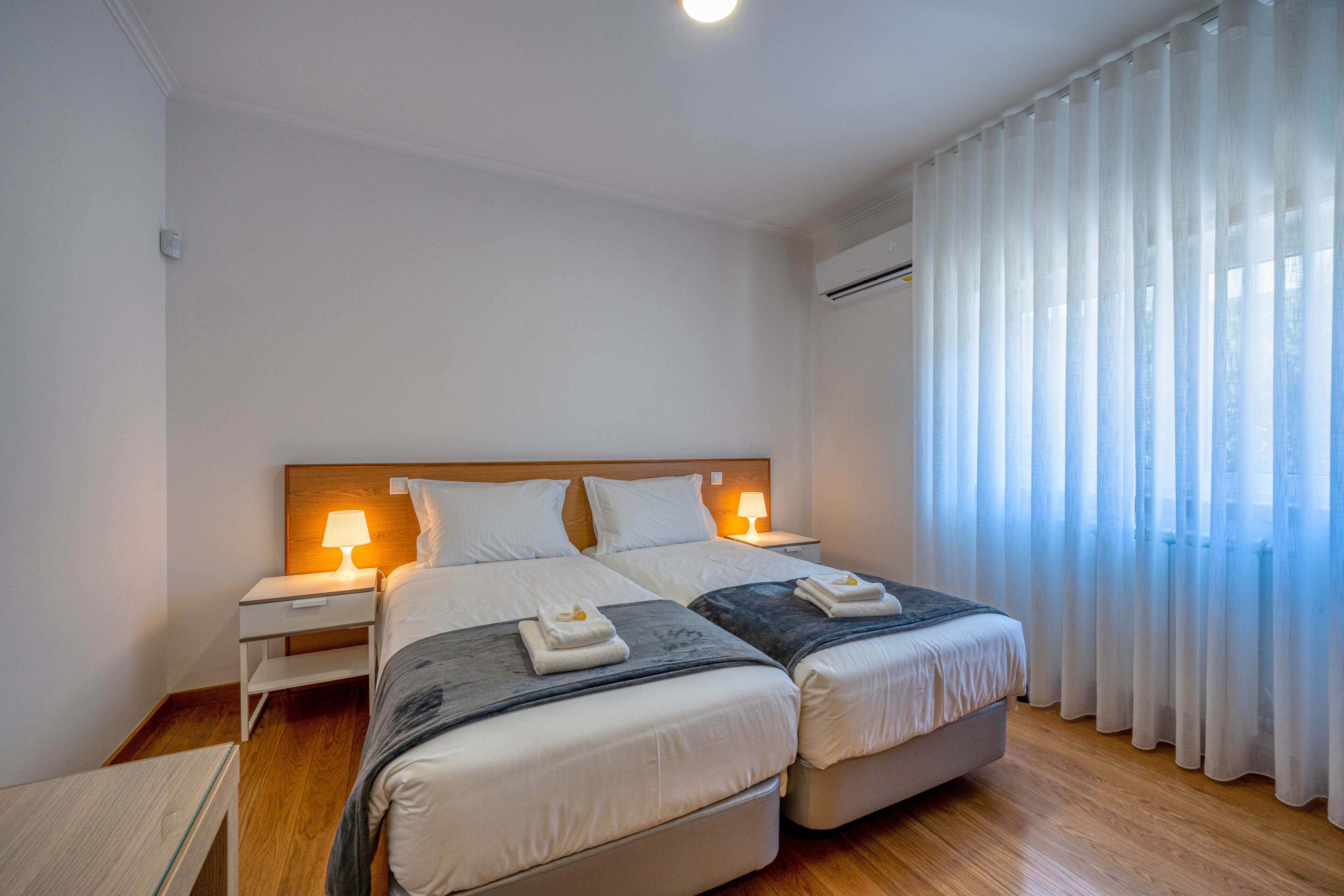 Habitación de hotel con dos camas, una mesita de noche con lámpara y una ventana con cortinas traslúcidas.