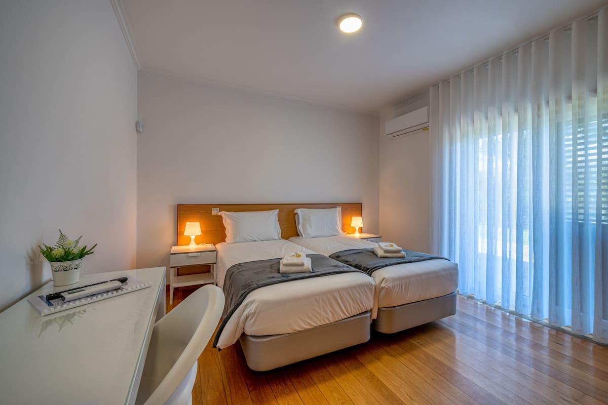 A hotel room with two beds, a desk, a chair, a potted plant, and a large window with curtains.