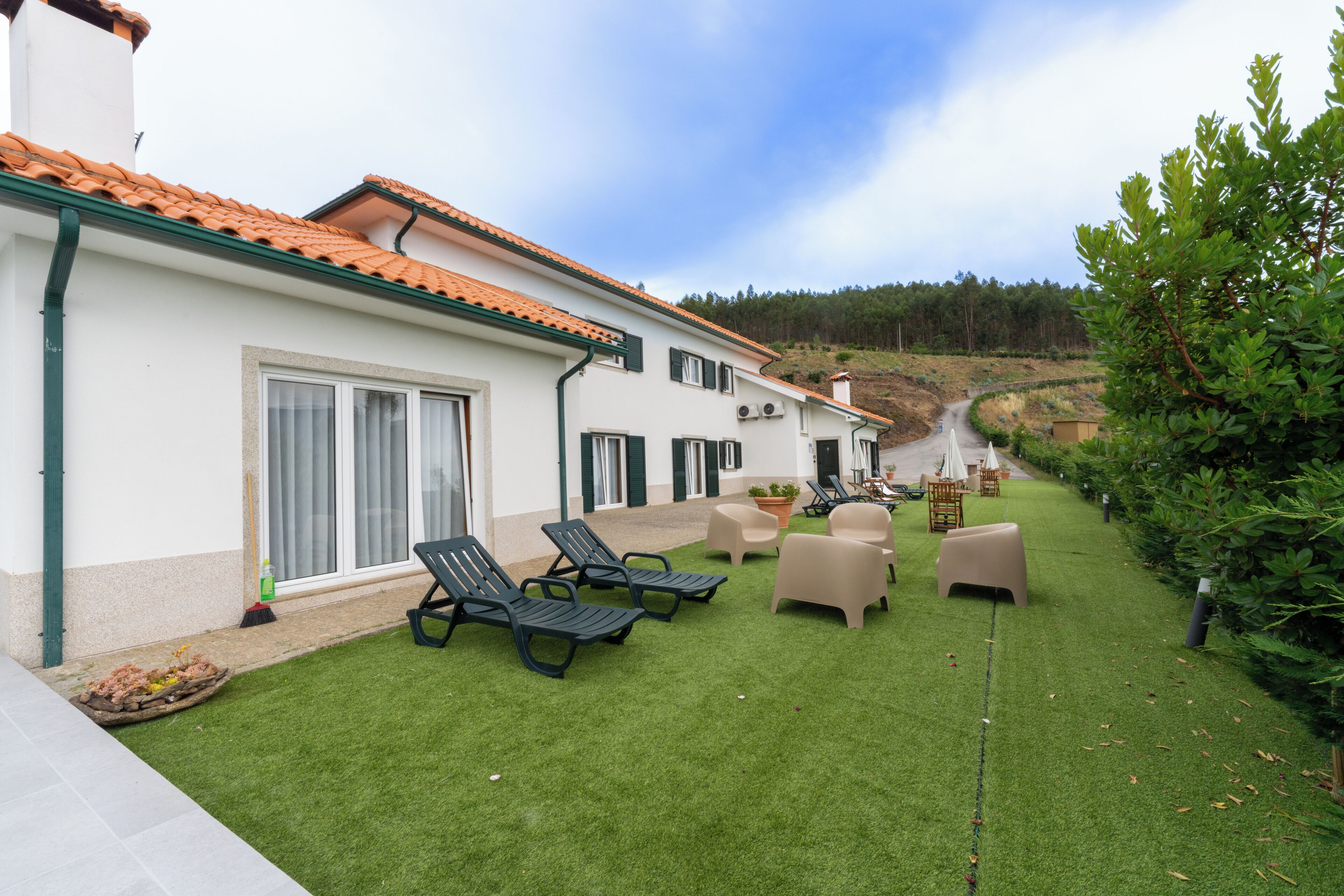 A two-story building with a tiled roof, a grassy patio with lounge chairs, and a pathway leading to a forested hill.