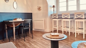 Un comedor con mesa de madera, dos sillas, una mesita redonda con un libro y una ventana con cortinas blancas.