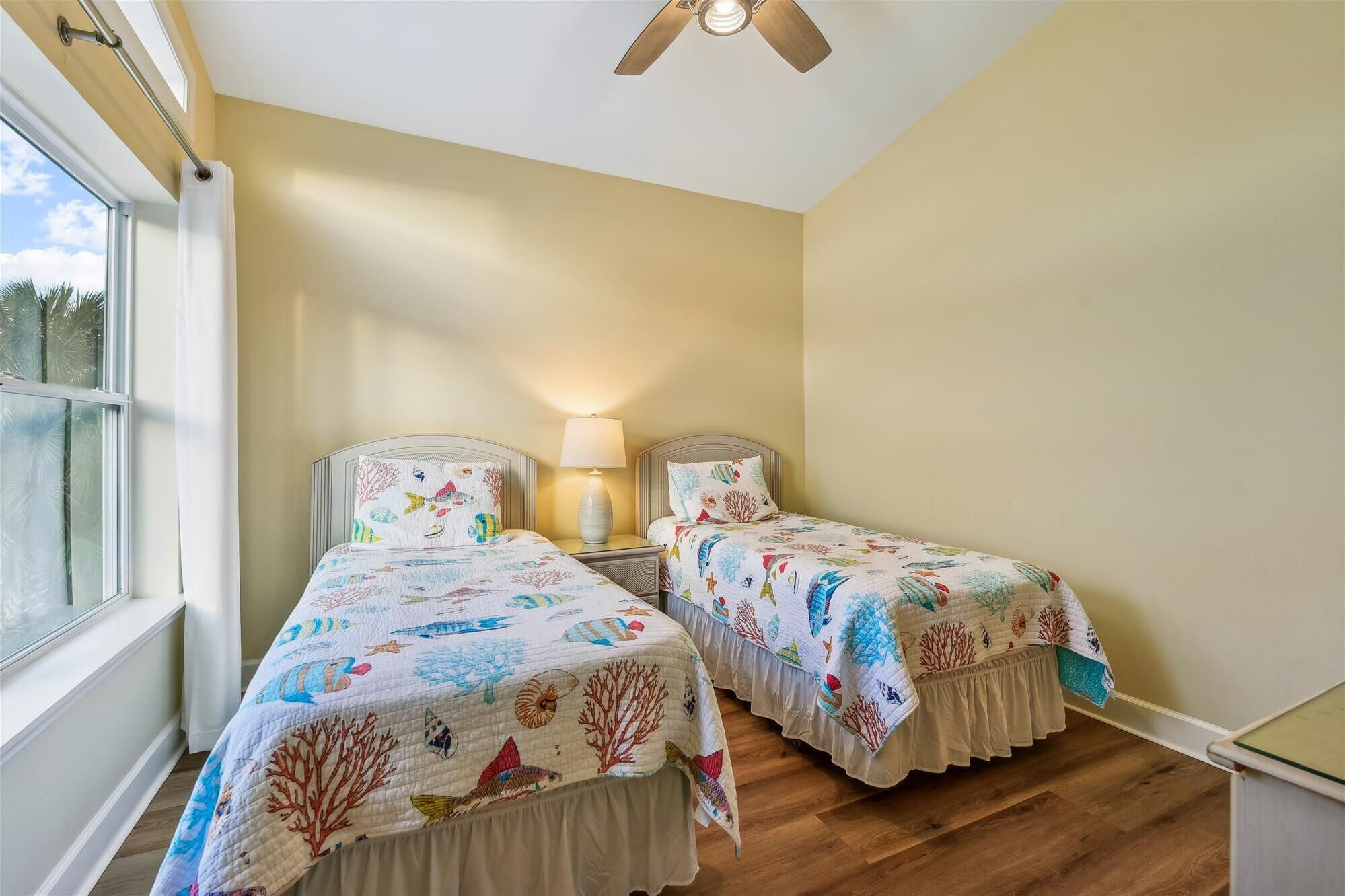 A room with two beds, each with a sea-themed bedspread, a ceiling fan, and a window with a view of palm trees.