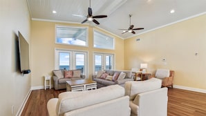 A spacious living room with a large window, ceiling fans, and a flat-screen TV mounted on the wall.