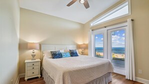 A bedroom with a bed, two bedside lamps, a nightstand, and a view of the beach through a sliding glass door.