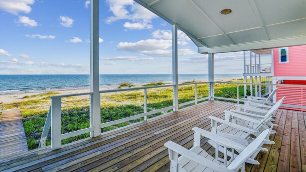 A covered wooden deck with white Adirondack chairs, overlooking a beach with a clear view of the ocean.
