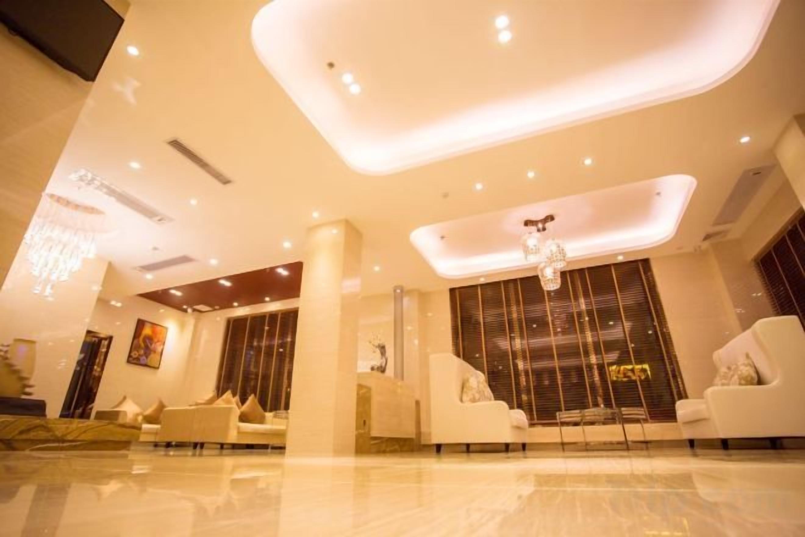 A modern hotel lobby with a high ceiling, recessed lighting, and a central chandelier.