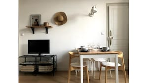 Un comedor con una mesa de madera, sillas blancas, un televisor sobre un soporte negro y un sombrero colgado en la pared.