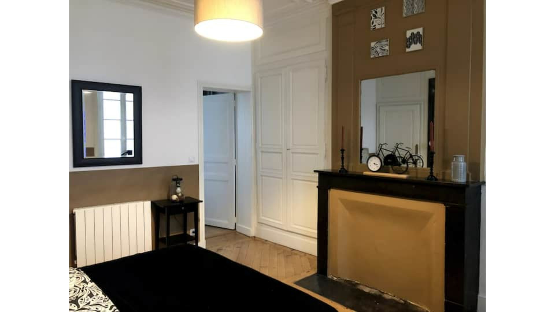A room with a black and white patterned bed, a white radiator, a small table with a lamp, a white door, and a mirror on the wall.