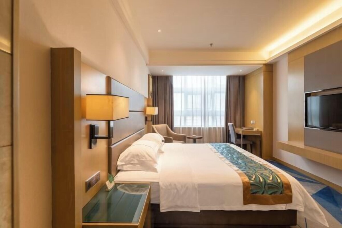A hotel room with a bed, bedside table, lamp, TV, and a view of the outdoors.