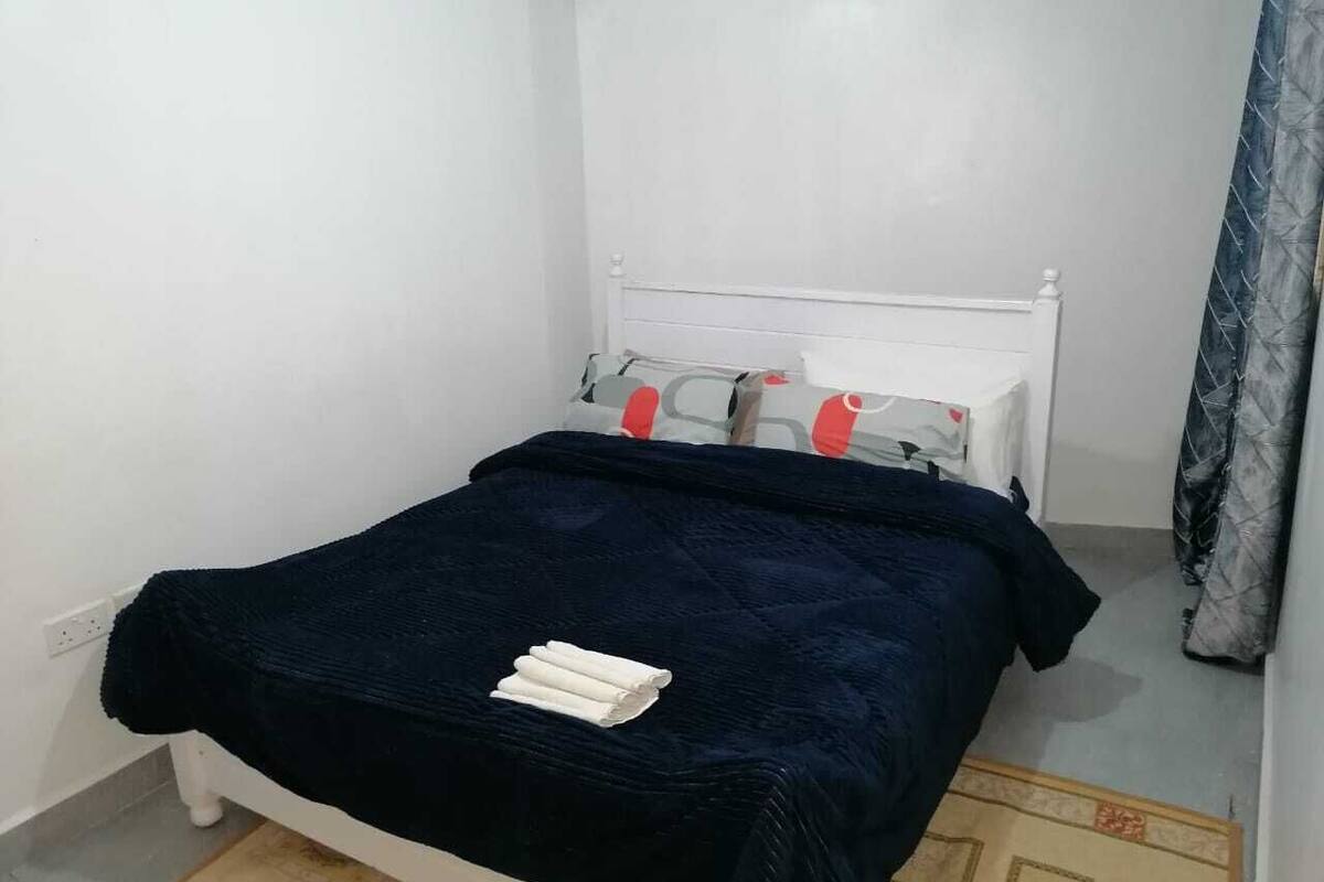 A small, neatly made bed with a dark blue bedspread and white pillows, a white headboard, and a blue curtain partially visible.