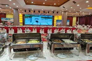 A buffet setup with multiple serving stations, a large screen displaying a scenic view, and a formal dining area with white chairs and red tablecloths.