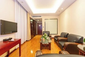 Ein Hotelzimmer mit Flachbildfernseher, einem Holztisch, einem Glastisch mit Blumen, Ledersofas und einem großen Fenster mit Jalousien.