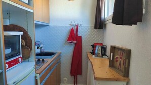 Cucina compatta con forno a microonde, tostapane e piccolo frigorifero. Si notano un pensile in legno e un piccolo piano di lavoro. Un asciugamano rosso è appeso al muro e sopra il lavello è montato un orologio.