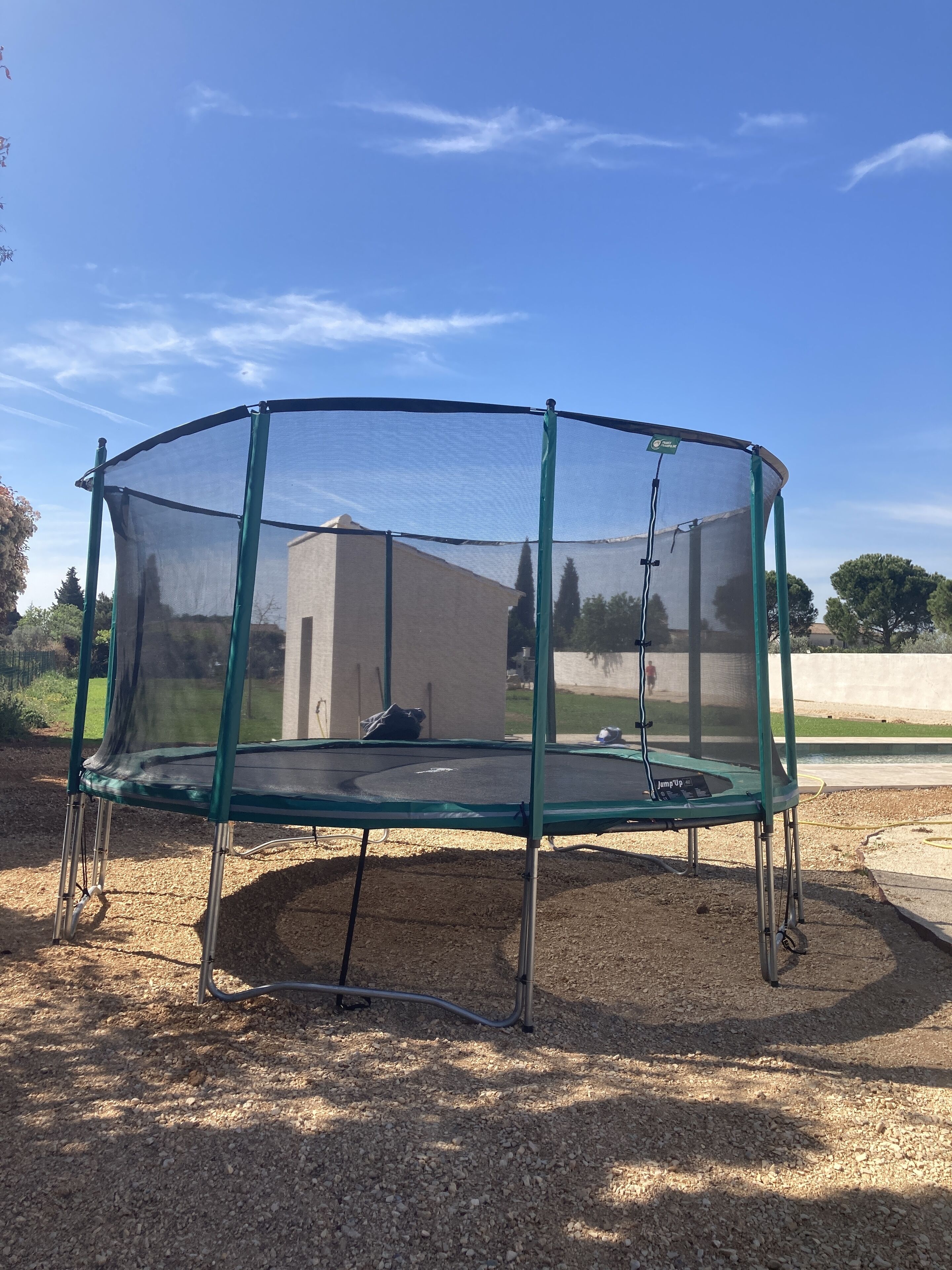 Un trampoline vert doté d’un filet de sécurité, installé sur une surface de gravier dans un espace extérieur.