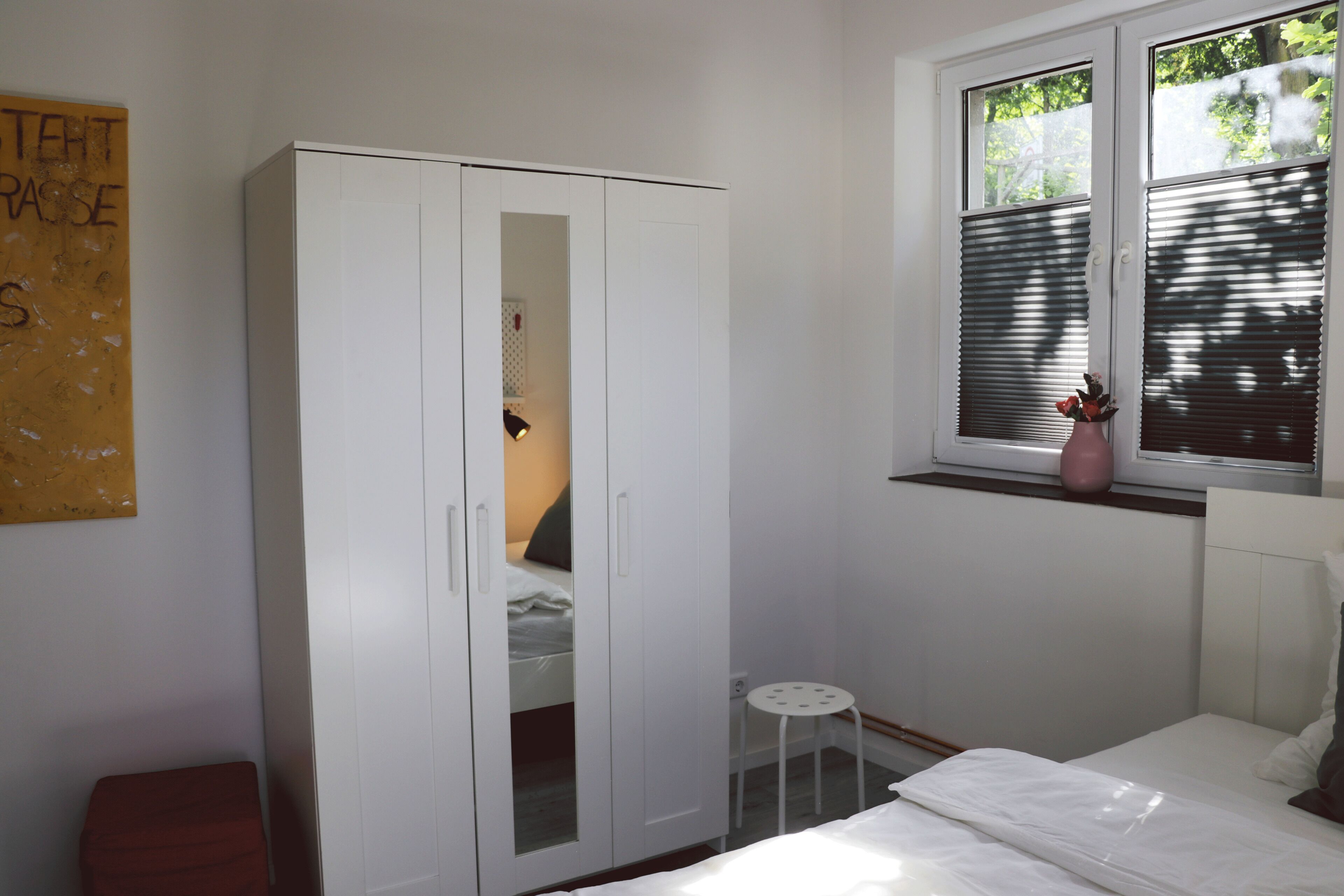 Quarto com guarda-roupa branco, cama, mesa pequena e janela com persianas.