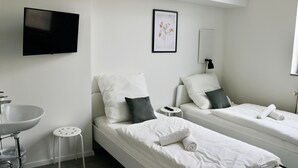 Una habitación de hotel moderna con dos camas individuales, un televisor de pantalla plana grande, un lavamanos blanco y una mesita redonda pequeña.