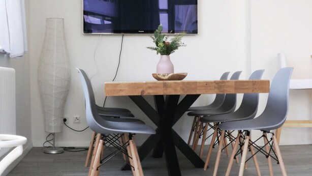 Un comedor moderno con una mesa de madera, sillas grises y una base negra. Un televisor de pantalla plana está montado en la pared sobre la mesa.